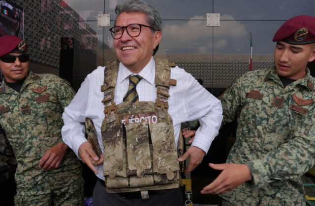 Ricardo Monreal prueba un chaleco táctico acompañado de dos militares durante una exhibición.
